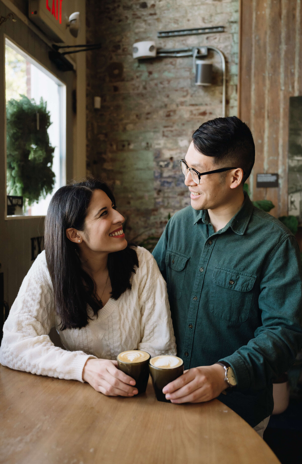 man standing next to a woman sitting at a wood table in a coffee shop with their cups together as they smile at one another for their engagement photos