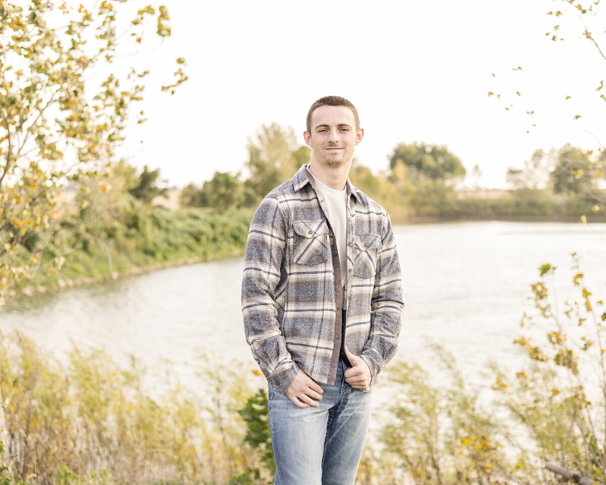 high school senior male standing in grass , with a pond and trees behind him, wearing a flannel jacket with a light tan shirt under, blue jeans, and looking at the camera with a soft smile