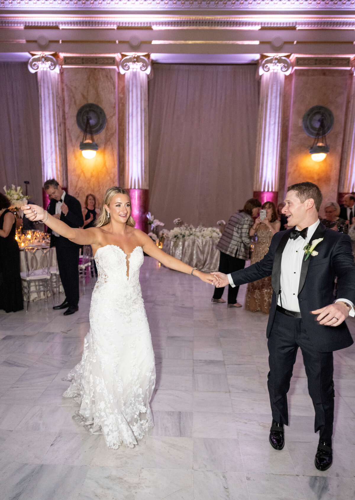 A bride and groom dance joyfully in a grand hall with elegant columns and soft purple lighting. She wears a strapless white gown, and he is in a black tuxedo. Guests watch in the background. Tables are adorned with floral centerpieces.