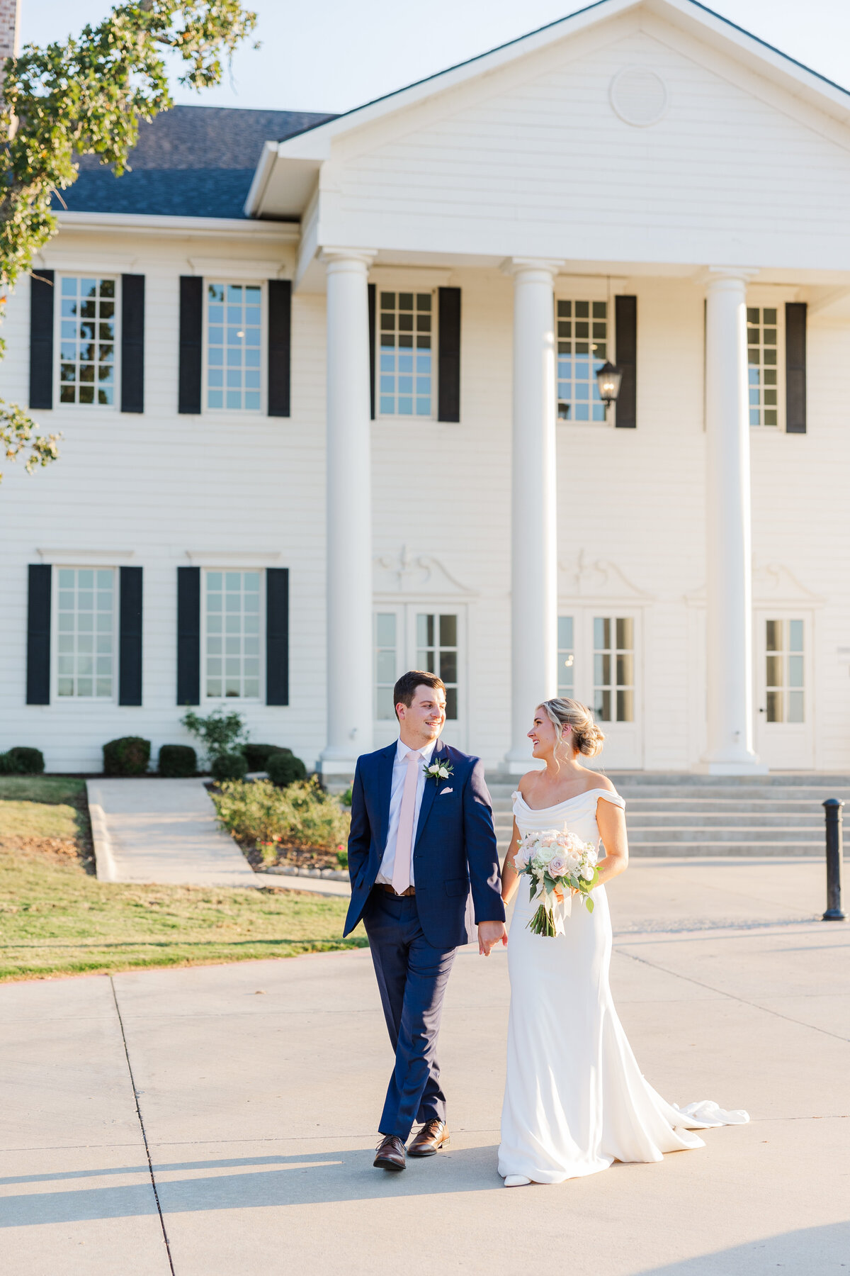 A bride and groom walk hand in hand outside a classic white-columned venue, bathed in golden evening light. Captured near Highland Village, this editorial-style wedding portrait exudes timeless elegance—ideal inspiration for couples planning a romantic wedding near Frisco or Flower Mound.