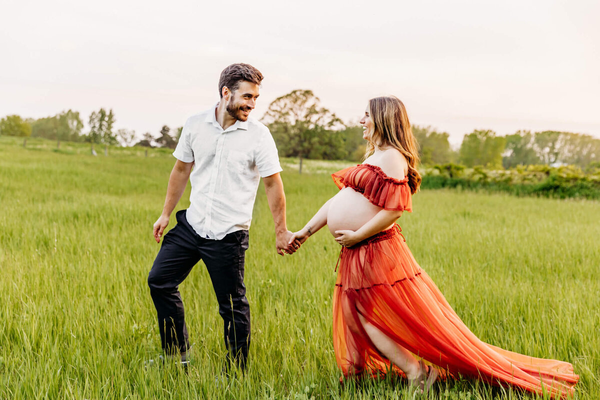 beautiful couple laughing as they walk together on a hill while the wife holds her baby bump captured by Green Bay Maternity Photographer Ashley Kalbus