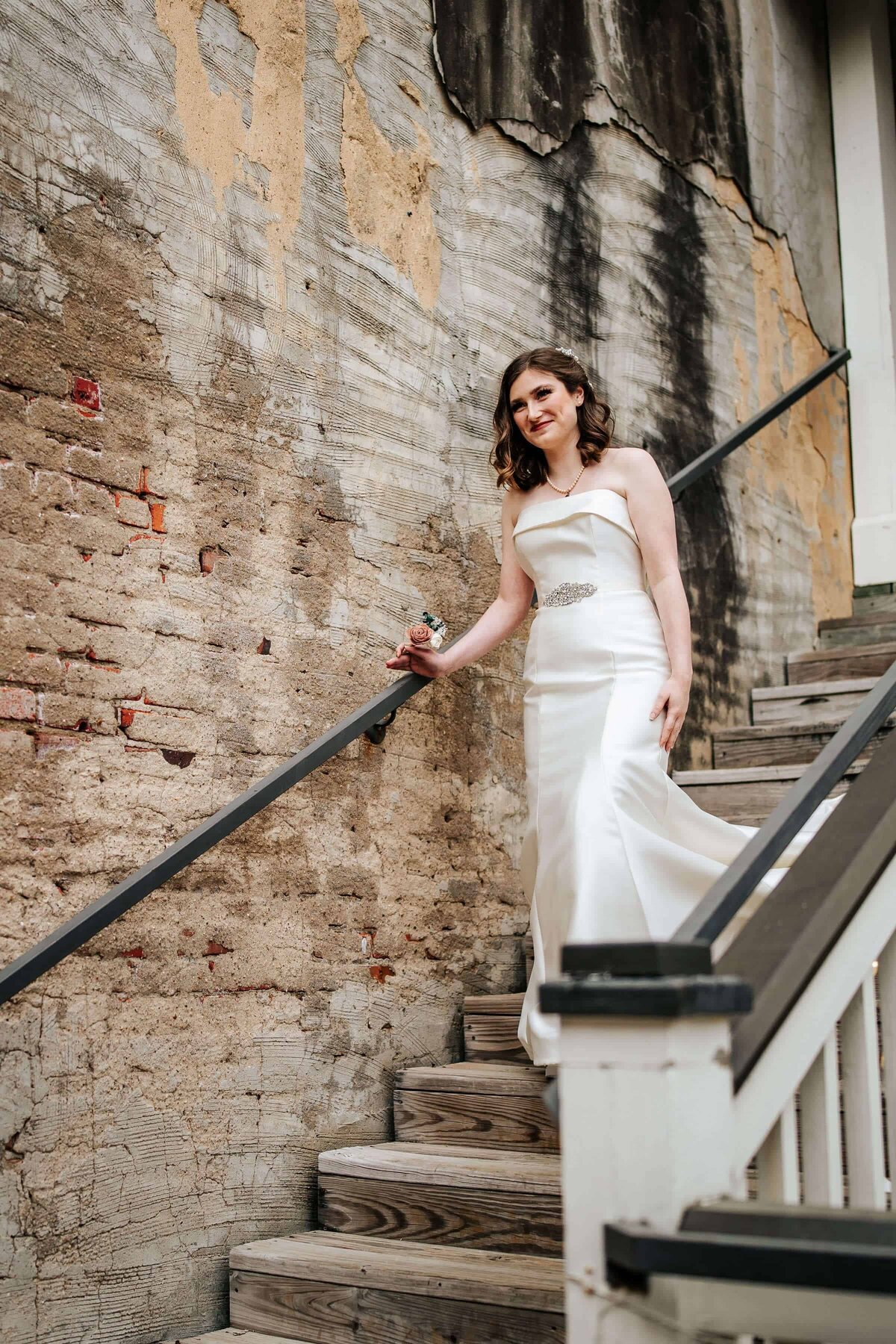 A bride in a white strapless gown with a jeweled belt walks down wooden stairs beside a weathered, textured wall, smiling and holding the stair rail.