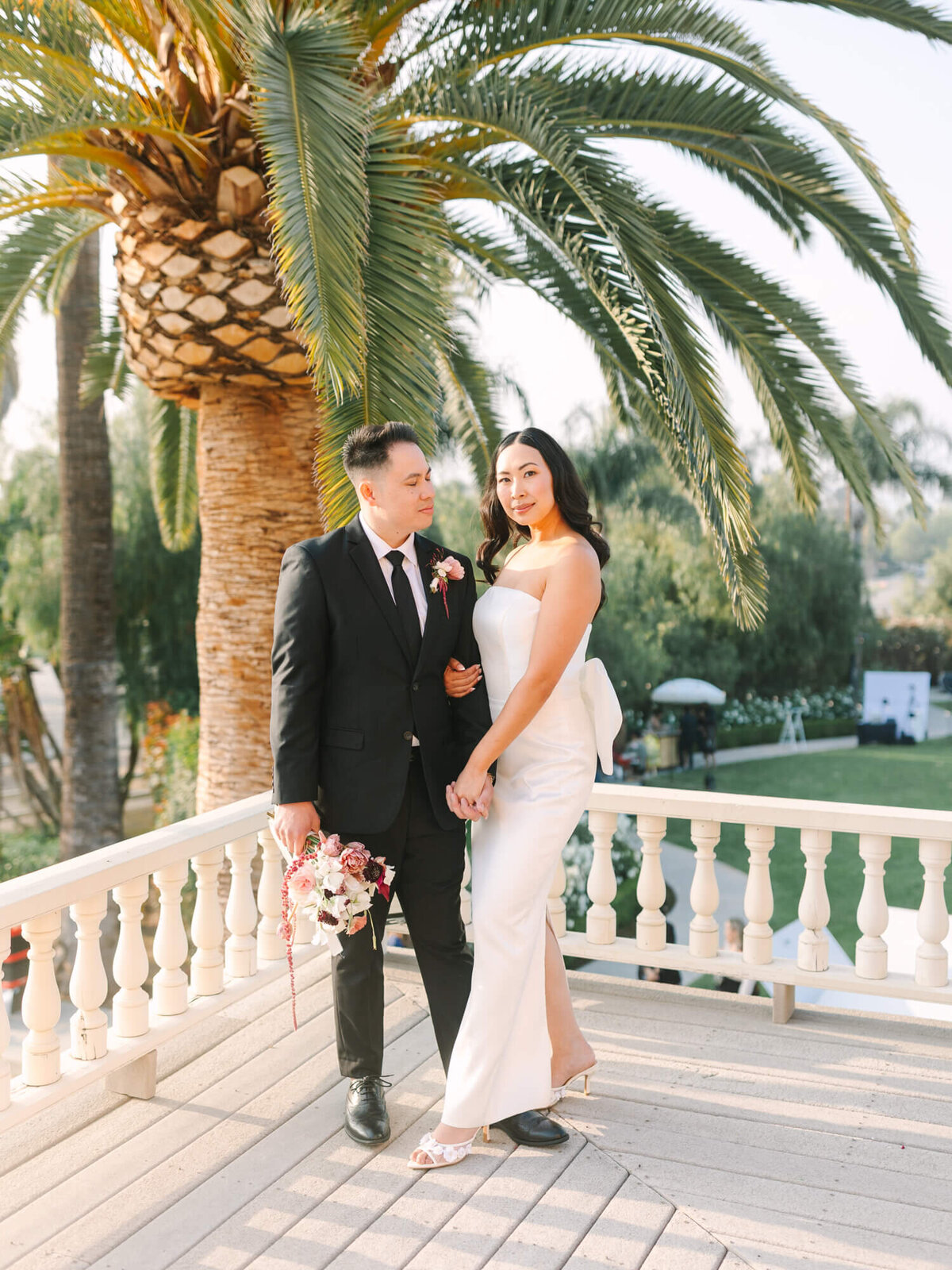 A couple stands on a sunny terrace, with a large palm tree behind them. The woman wears a sleek white dress and the man in a black suit.