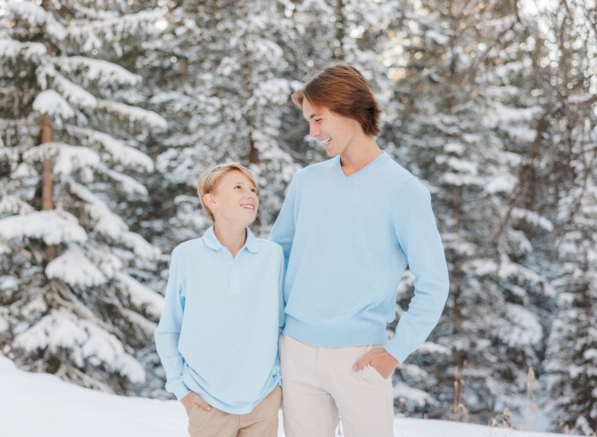 Teen brothers in blue shirts hug while smiling in a snowy forest
