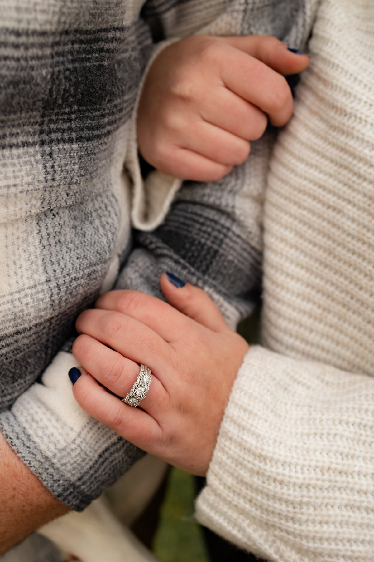 In a scene fit for a New England wedding, two individuals in cozy sweaters hold hands. One wears a gray plaid sweater, the other a white knitted one with dark blue nail polish and a sparkling diamond ring—a perfect moment for any New England wedding photographer to capture.