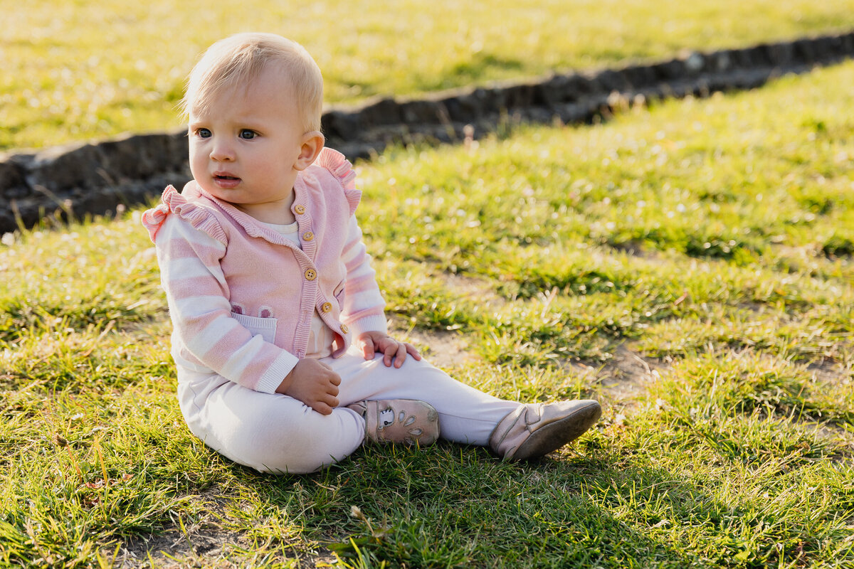 A baby wearing a pink striped jacket and white pants sits on grass, looking to the side with a curious expression.