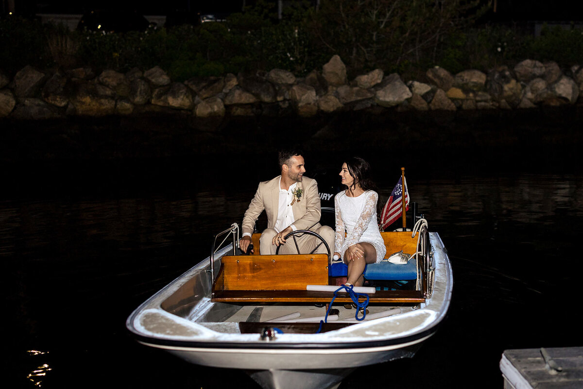 The bride and groom enjoy a quiet, intimate moment in a boat on the water at night, with lights reflecting on the water.