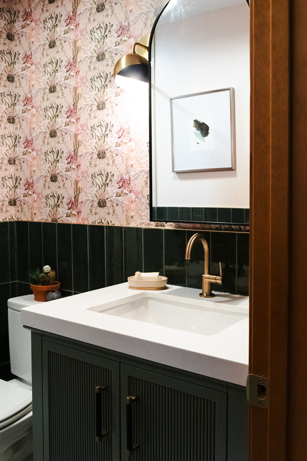 A stylish bathroom detail featuring a dark green vanity with vertical paneling and brass hardware, topped with a white rectangular sink and a curved brass faucet. A bar of soap rests on a small dish next to the sink. Above the vanity, a partially visible arched mirror reflects a framed abstract artwork on a white wall. The walls are decorated with dark green vertical tiles on the lower half and floral patterned wallpaper in shades of pink and beige on the upper half. A brass gooseneck wall lamp illuminates the vanity area. A glimpse of a toilet with a small potted succulent on its tank is visible to the left. A portion of a wooden door frame is on the right.