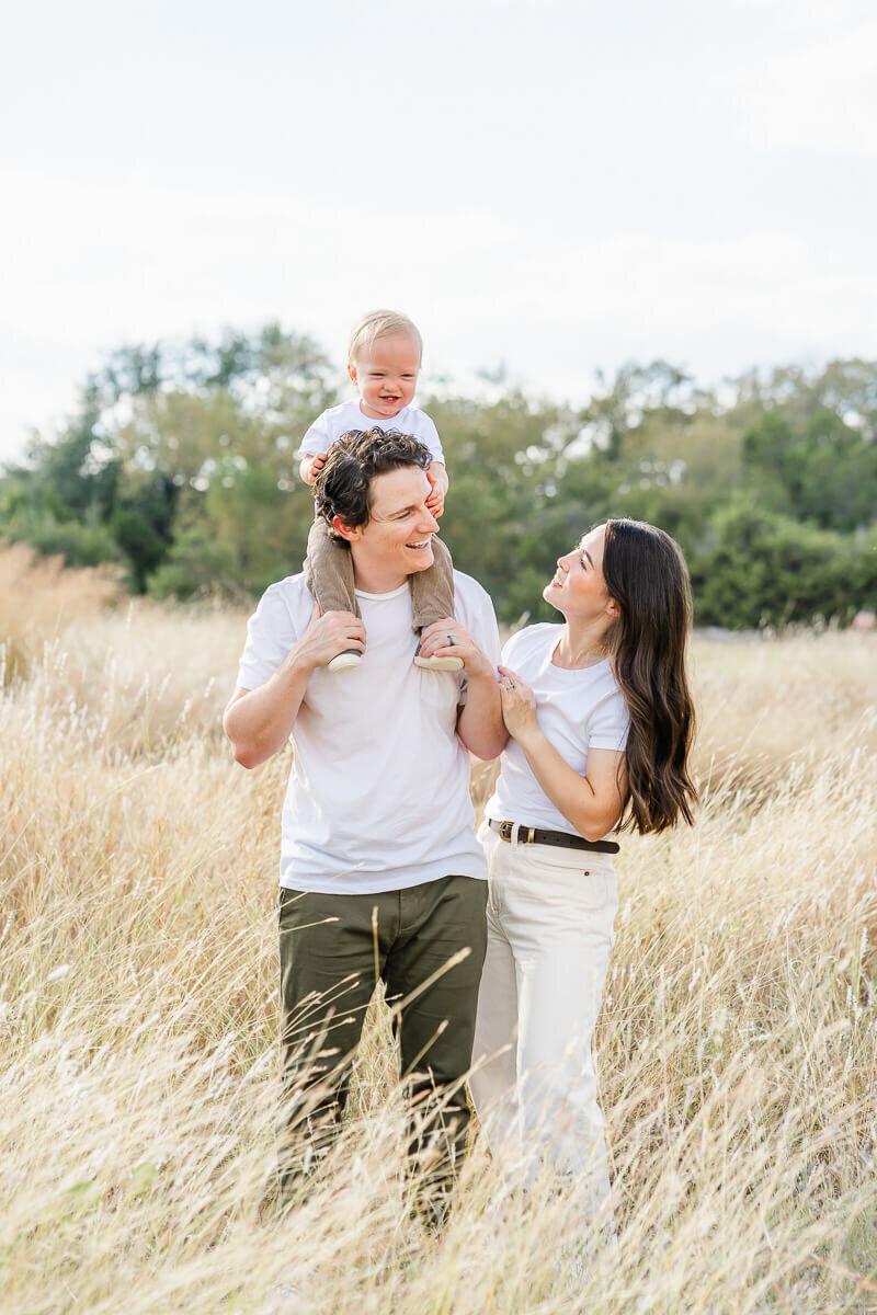 a mother and father look at each other with a smile while their toddler son sits on his father's shoulders for their fall family portraits.