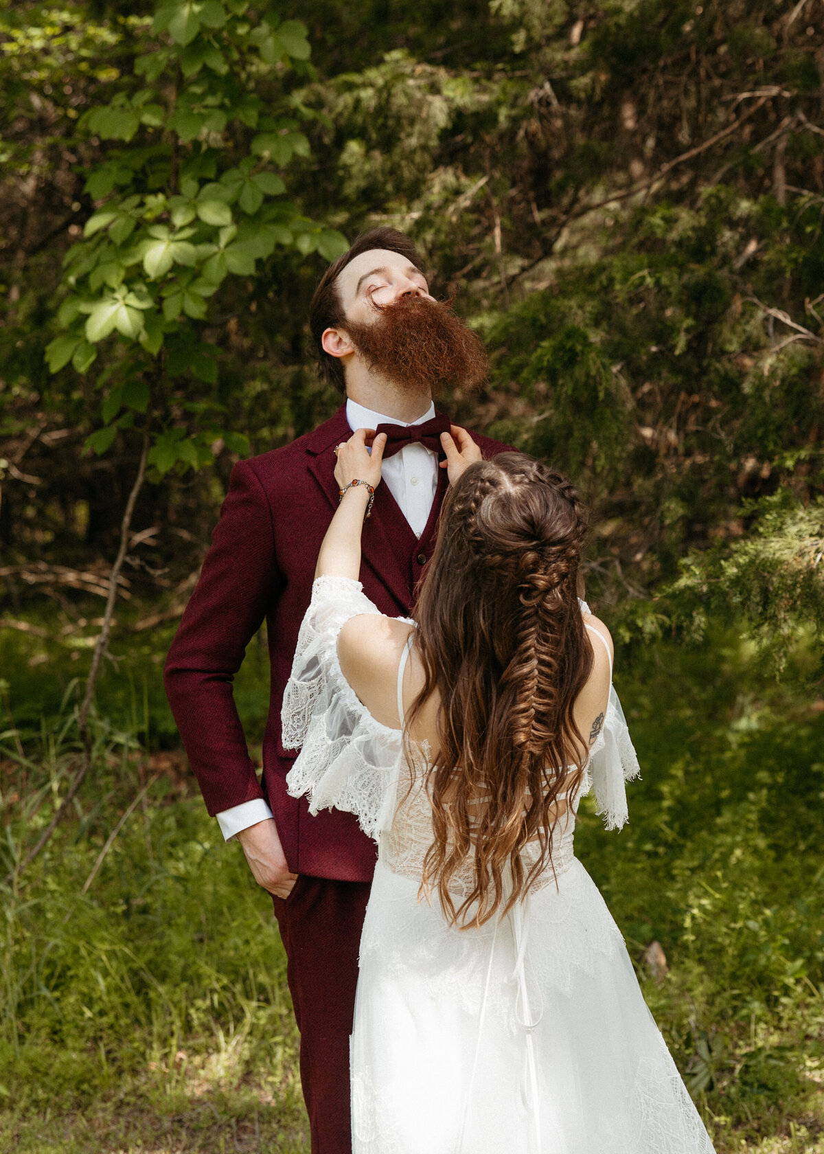 Bride playfully adjusting the bow tie of her groom who is clad in a maroon suit, both surrounded by lush greenery