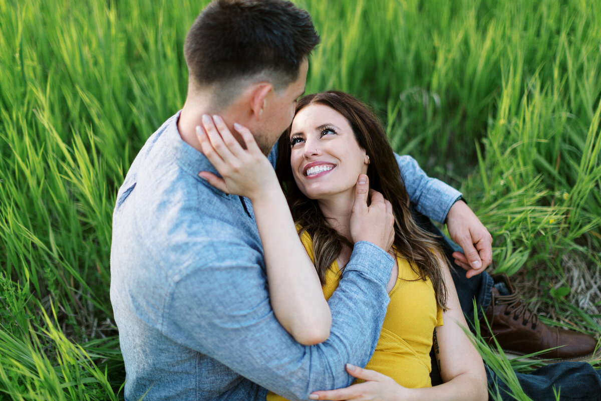 Couple sitting in the tall grass of Maple Grove and looking at each other with smiles.