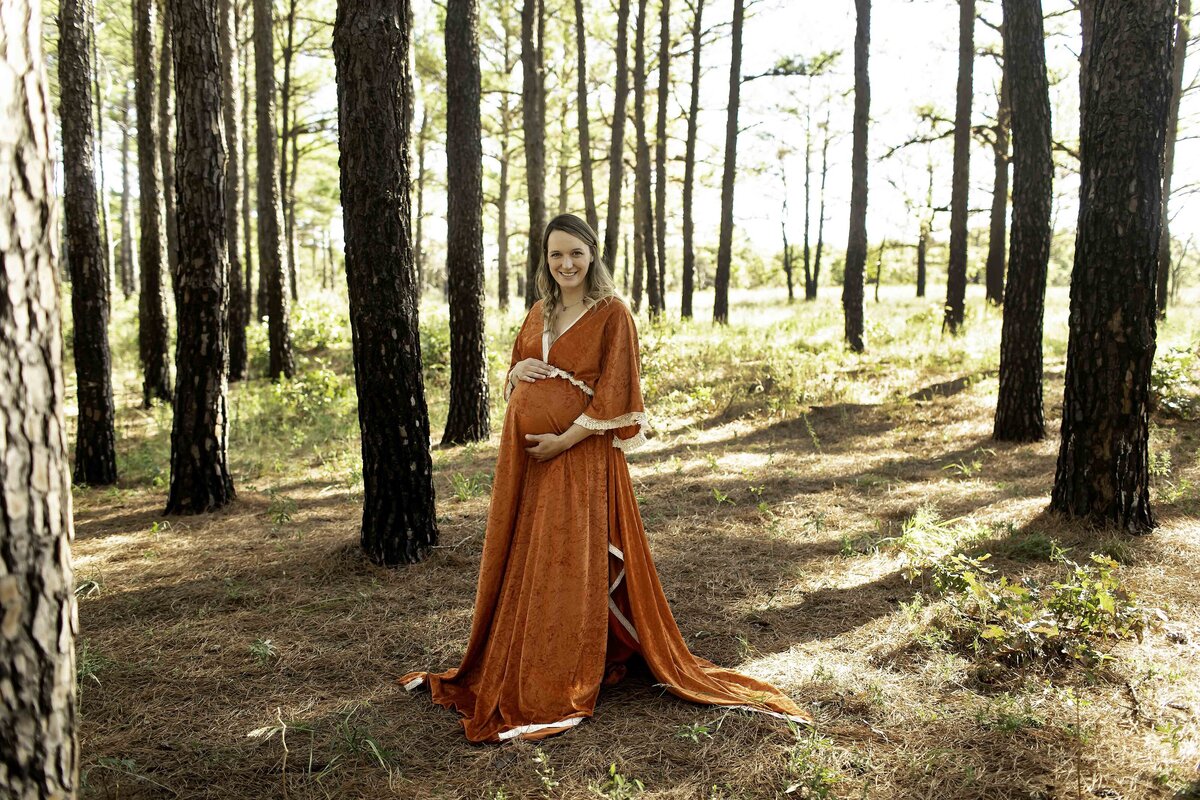 A tranquil maternity photo of the expectant mom standing in a lush pine tree forest, her hands lovingly resting on her belly, with sunlight peeking through the trees, beautifully captured by Chunky Monkey Photography.