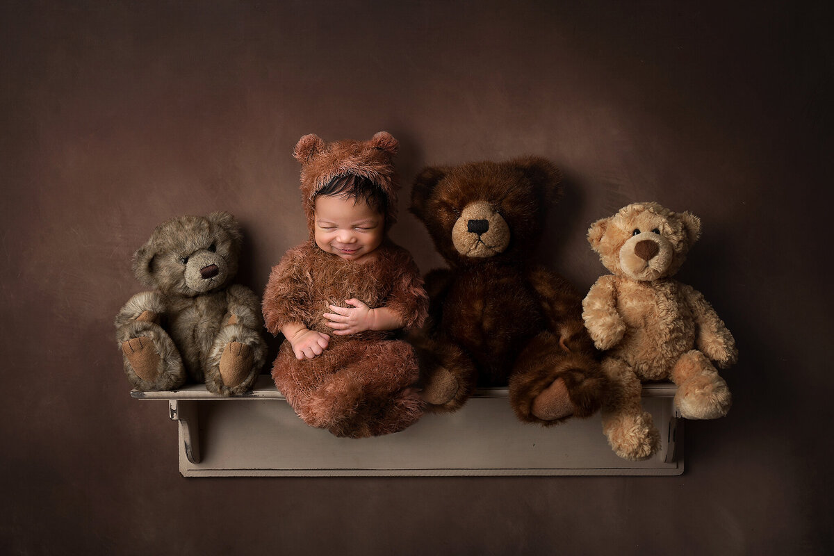 little newborn boy in a bear outfit posed on a shelf with teddy bears, brown tones on a brown textured background