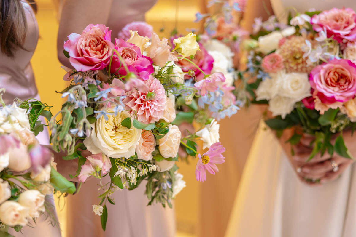 Three people in pastel gowns hold vibrant flower bouquets featuring pink and white roses, peach dahlias, and assorted greenery. The bouquets are richly textured and colorful, adding a touch of elegance to the scene.