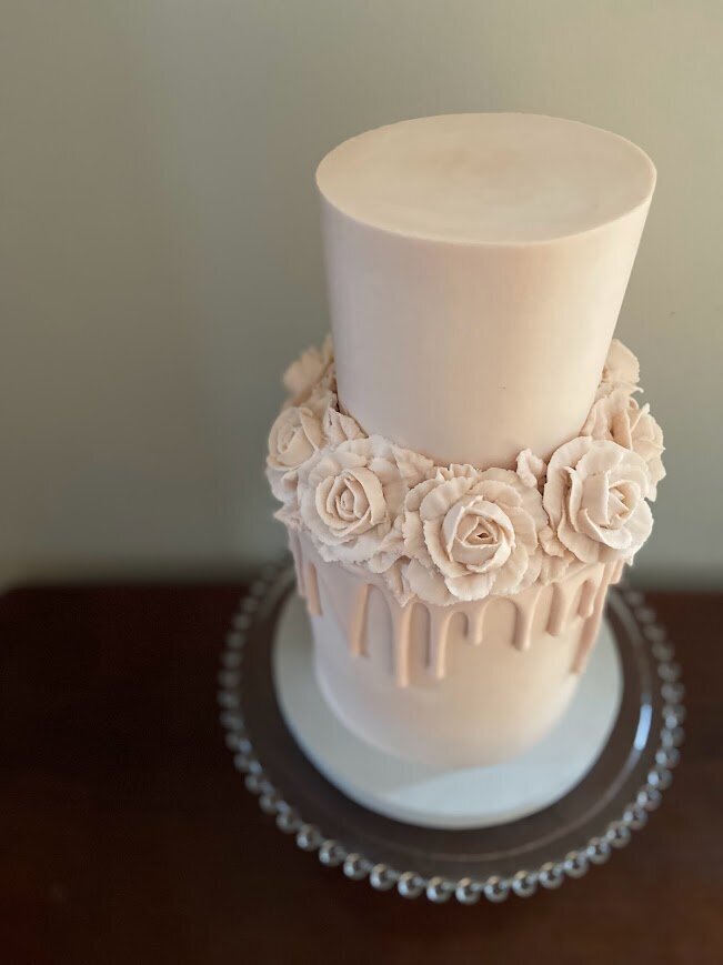 Photo shows are two tier pastel pink cake. The cake has buttercream flowers around the middle where the two tiers meet, in the same color as the cake is frosted. There is also a drip in a matching shade of pink, below the flowers.