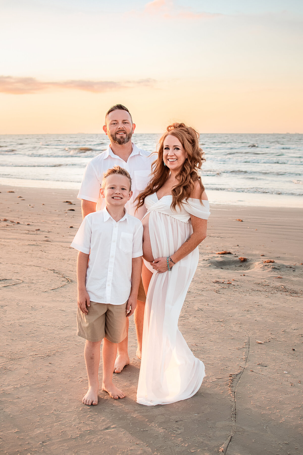 A mom, her husband, and son stand close together on a Galveston beach and smile at the camera.