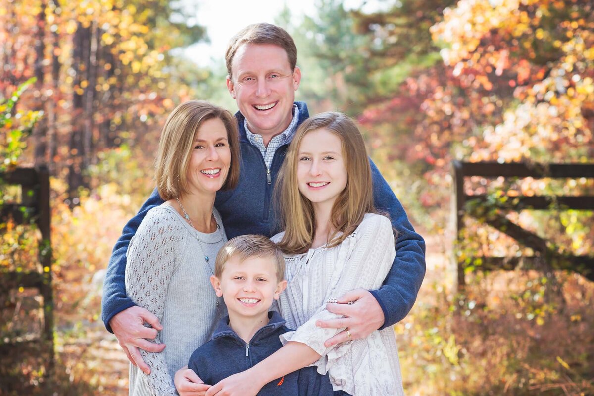 Family portrait with dad hugging his wife and children in a fall setting with a fence.