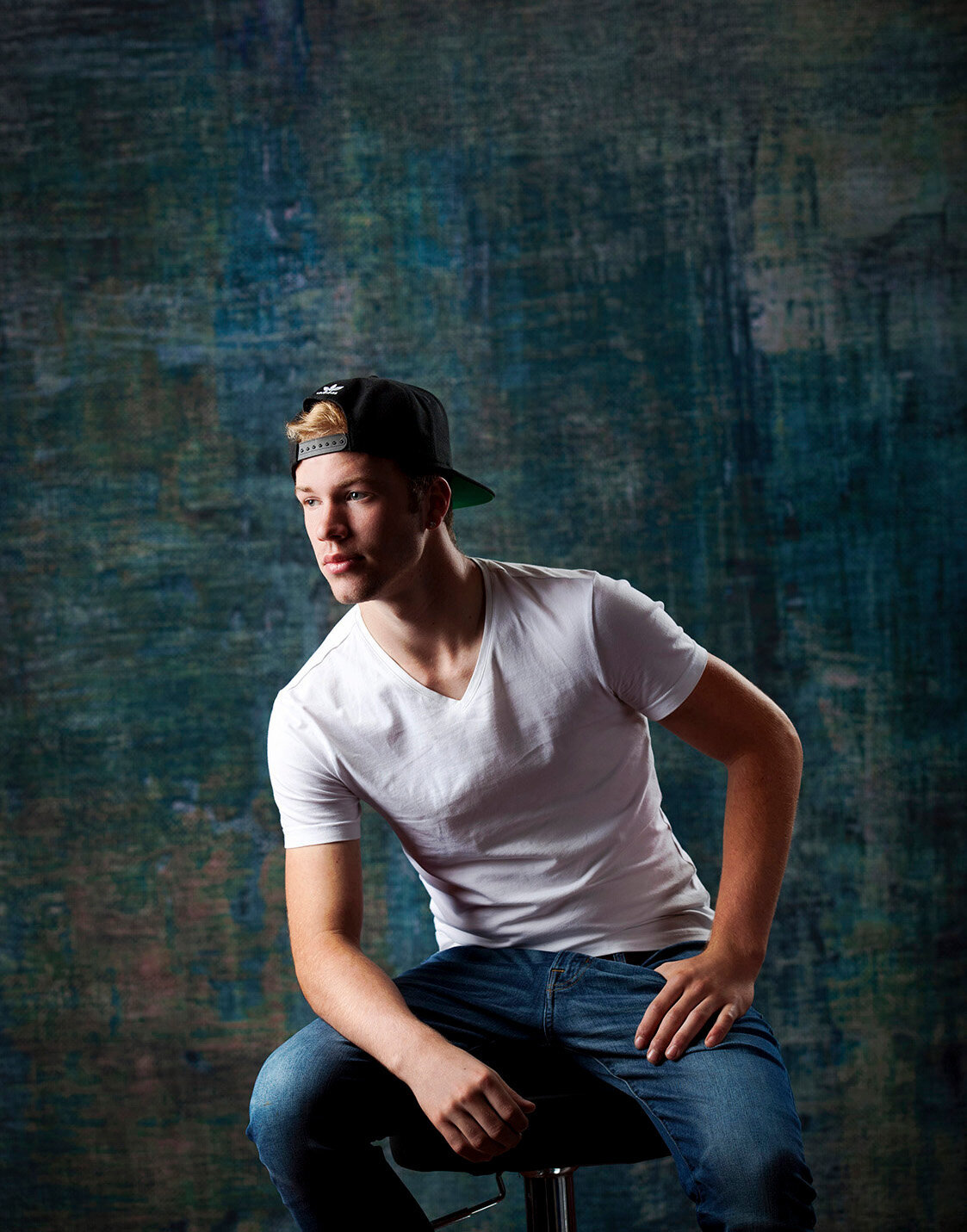 Young guy sitting on stool for studio portrait with baseball cap on backwards looking off to the side Studio 64 Photography Akeley Minnesota.