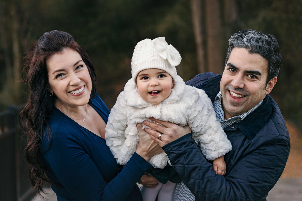 mom and dad swinging their little girl wearing fuzzy white winter clothes toward the camera in the winter