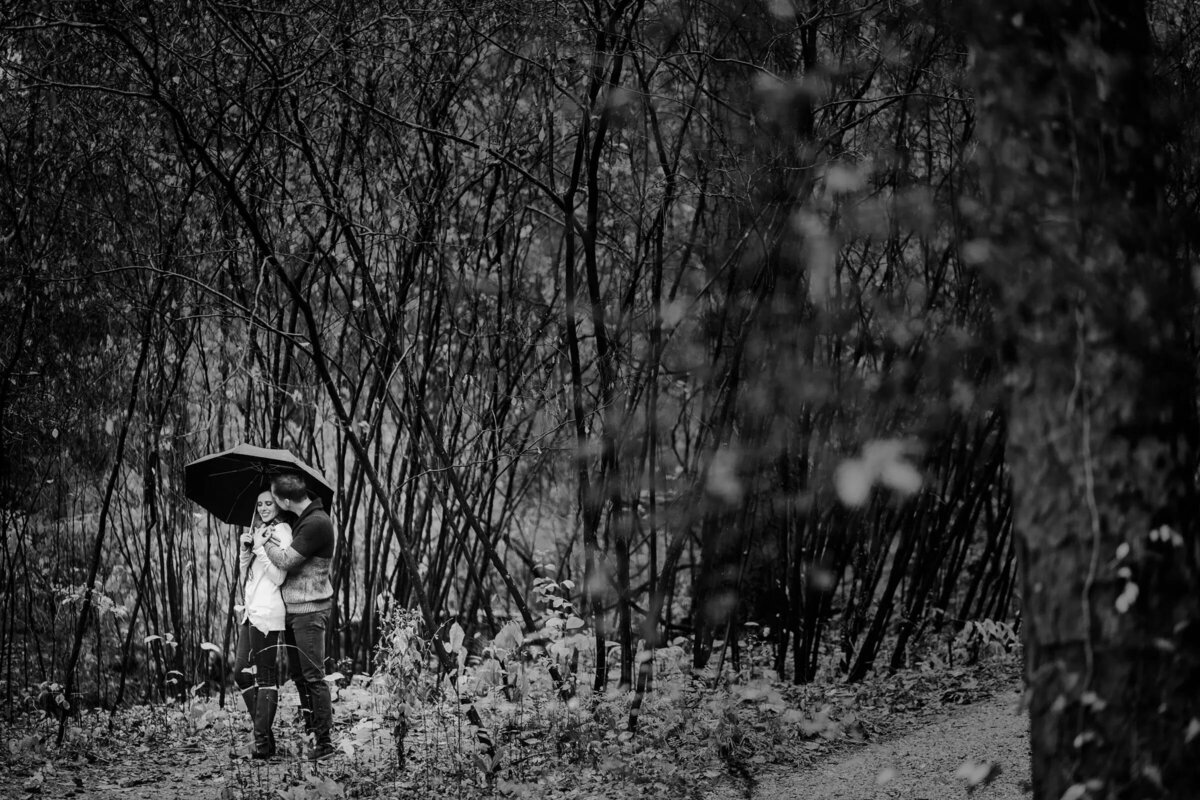 Monochrome photo of a couple sharing an umbrella, surrounded by the soft focus of a barren forest