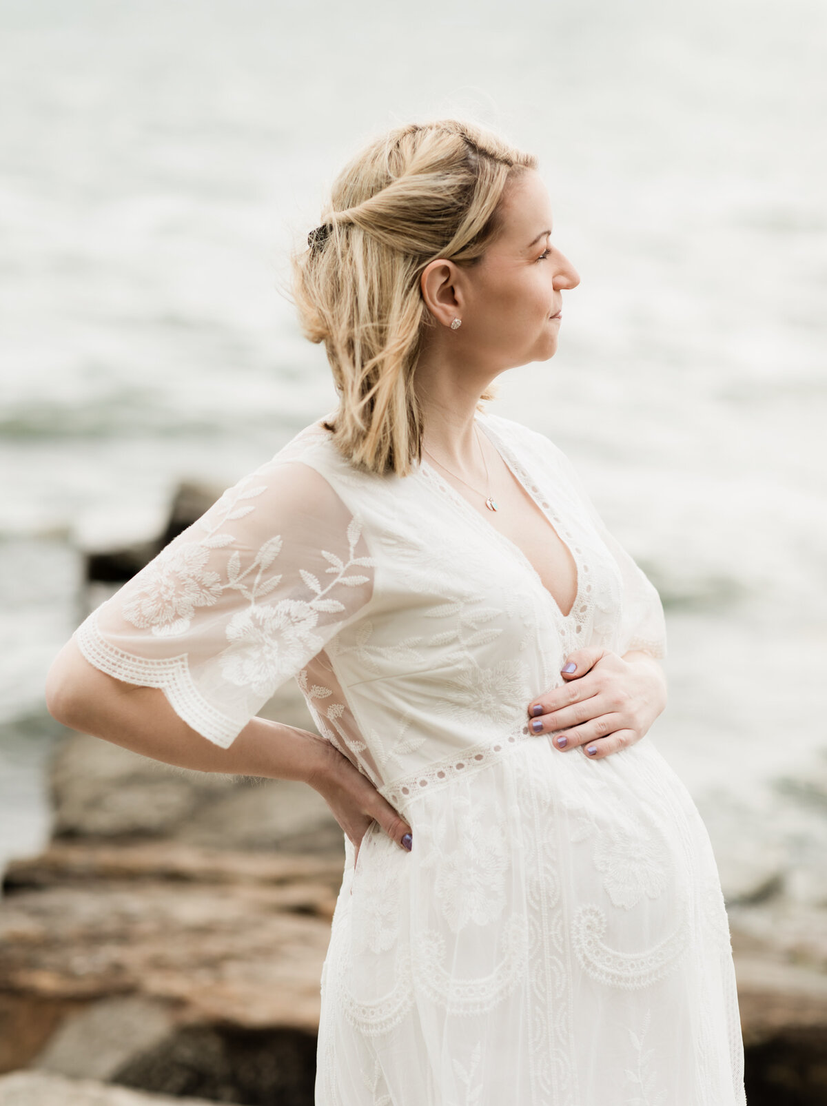 pregnant mother in white maternity gown holding baby bump on beach for maternity photoshoot 