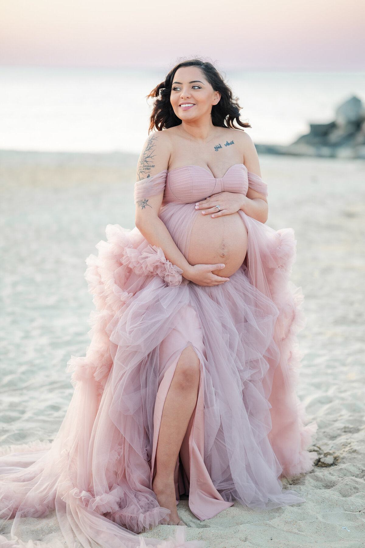 A mom-to-be holds her belly and smiles off into the distance. She is wearing a pink tulle gown at her beach maternity session.