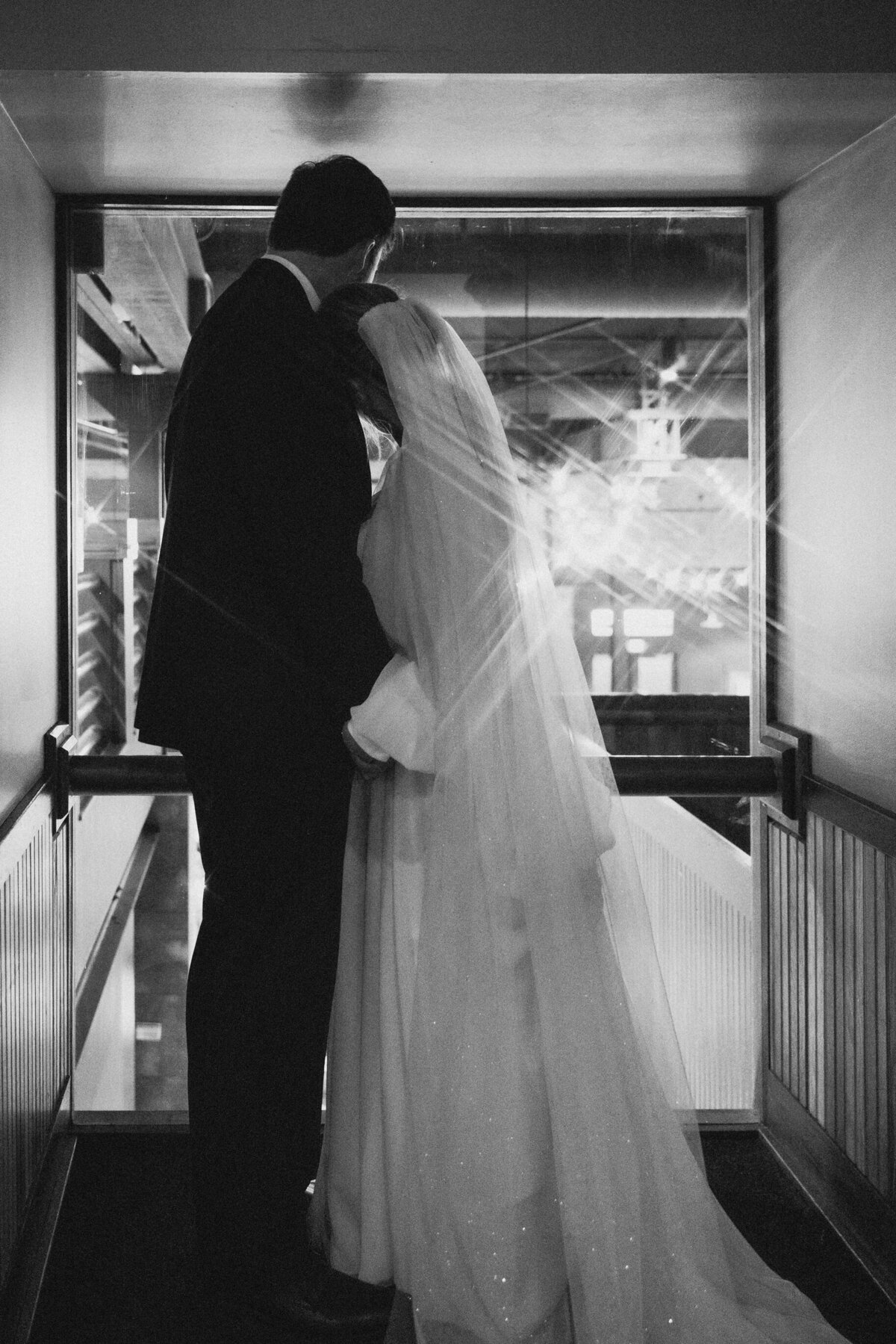 black and white of bride and groom leaning together as they overlook their reception from the top floor featuring a starburst in the corner of the image