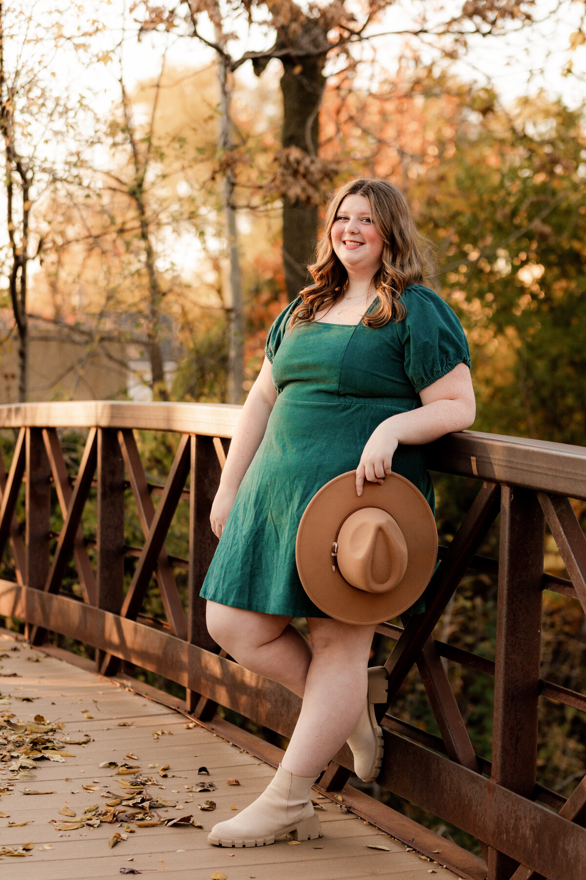 Claire holds her hat by her side while she leans against a brown bridge in a green dress.