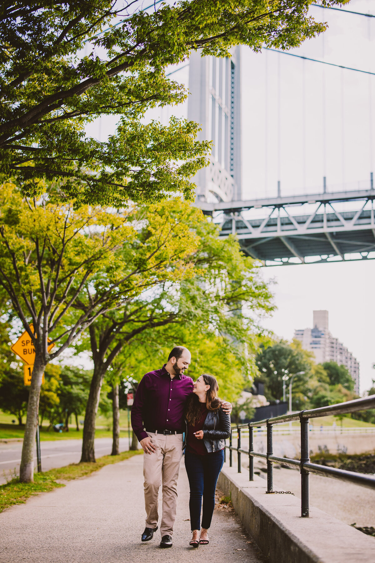 An engaged couple walking down the street at astoria park with the RFK bridge in the background