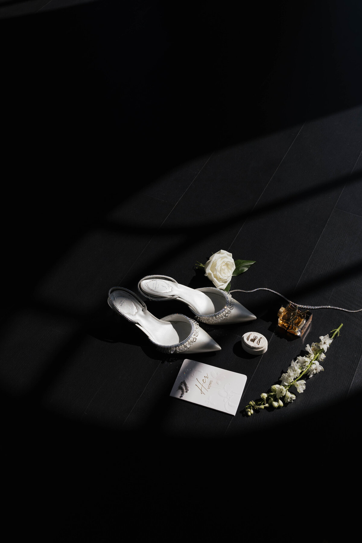 A luxury bridal flat lay featuring pearl-embellished heels, a white rose, elegant wedding rings, and a vow booklet in soft light. A refined and cinematic wedding detail shot.