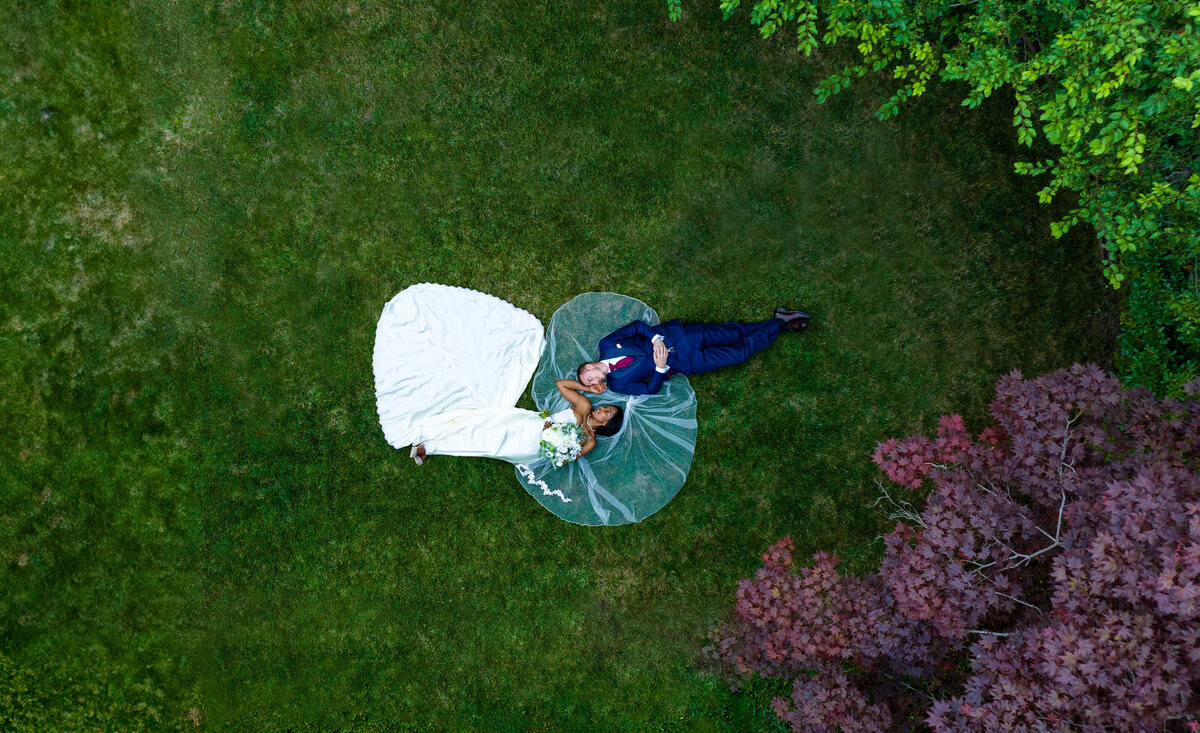 Bride and groom lay in a field with trees on their righthand side.  They lay opposite with their heads together looking at each other. Bride's dress and veil is spread out. Photo captured by drone above by wedding photographer, philippe studio pro.