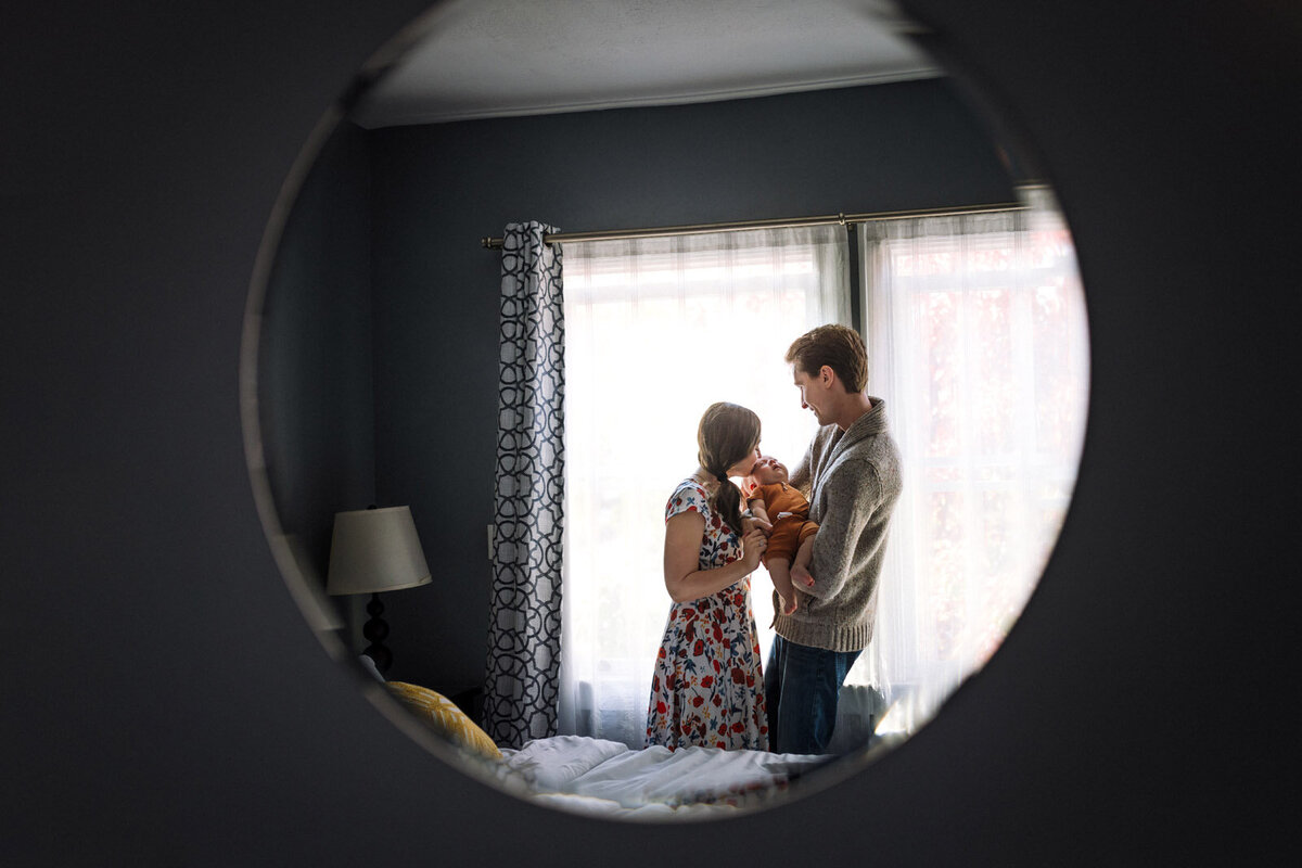 reflection in mirror of parents standing in front of window and mom kissing baby in dad's arms