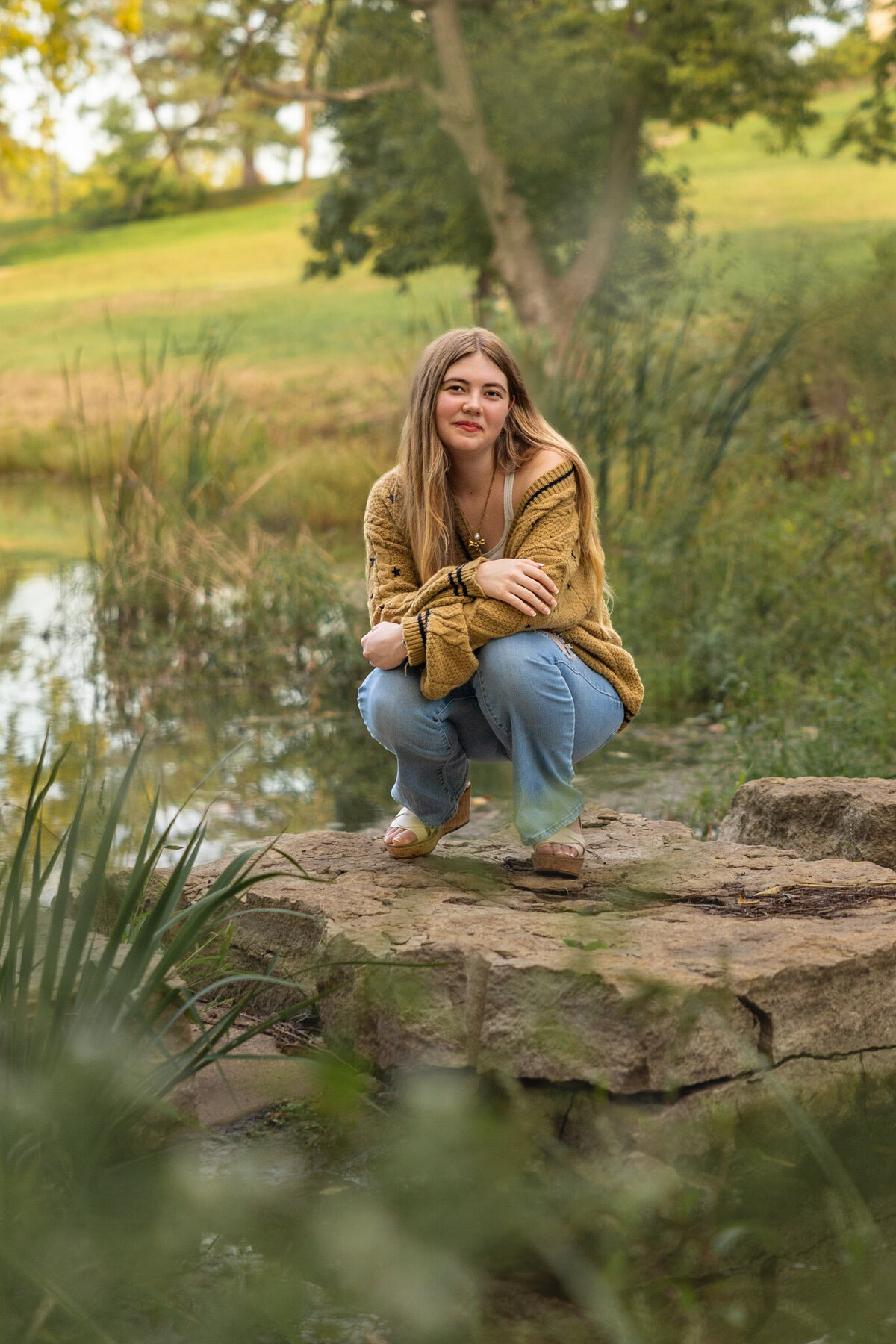 A senior girl squatting on a rock in the middle of a pond on the KU Campus in Lawrence, KS