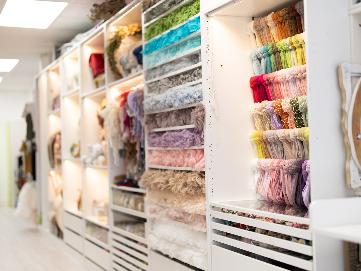 A neatly organized shelf displays folded, colorful textured blankets in pastel and bright hues. Soft lighting and adjacent hanging black garments add elegance.