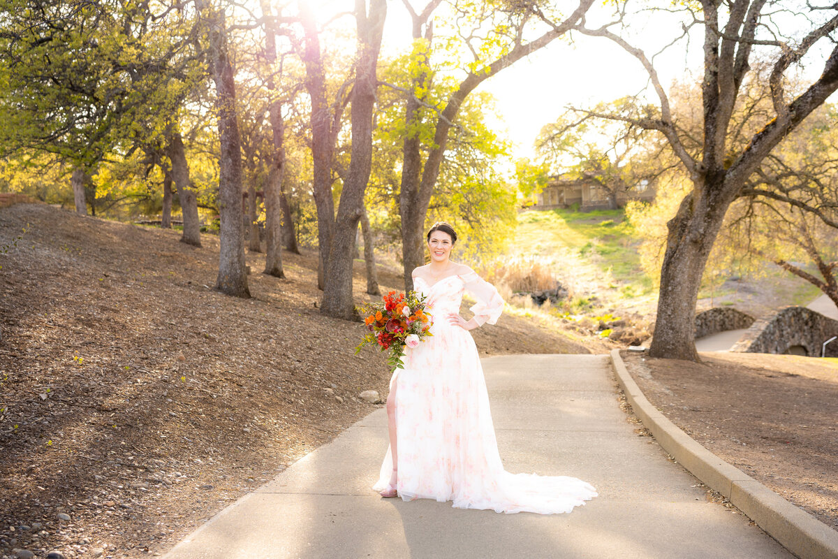 Bride stands on pathway in boho dress with pink and white pattern holding bouquet. She has her hands on her hips, a popped leg and is smiling at the camera with trees and sunset in her background. Photo captured by philippe studio pro, wedding photographer from Sacramento.