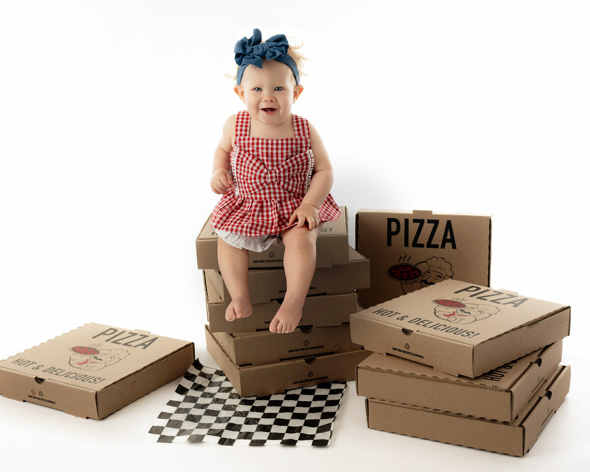 Child in red gingham dress sitting on pizza boxes with a blue headband.