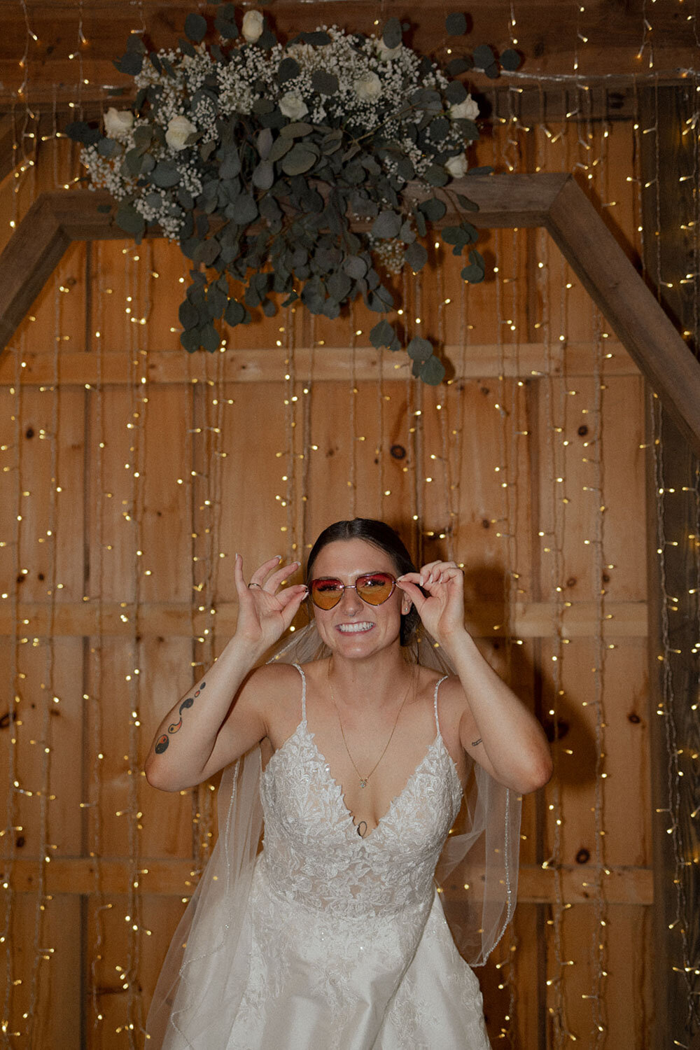 Bride smiling with sunglasses on, standing in front of a backdrop of twinkling lights