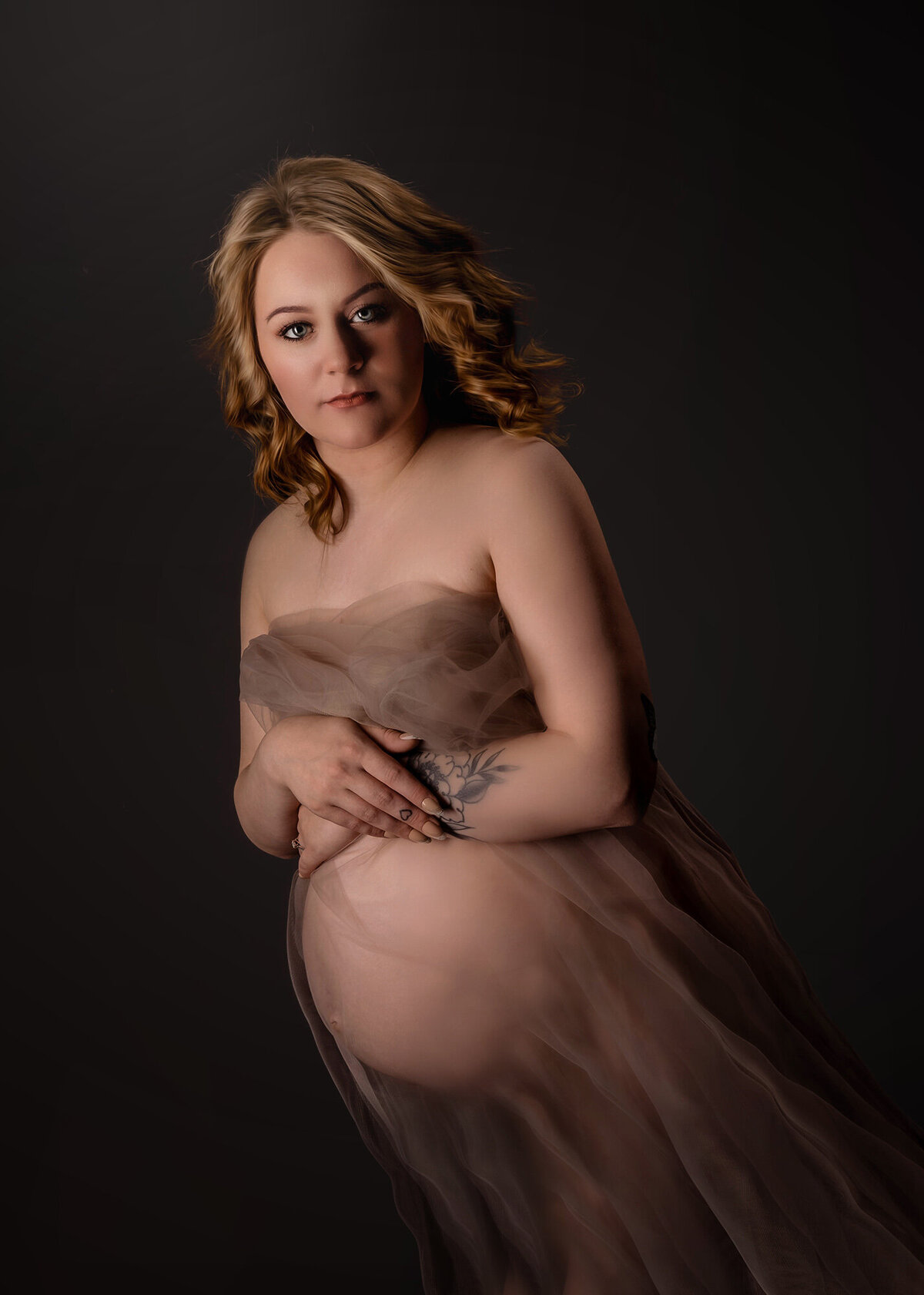 A mom to be stands in a studio covering herself with a sheer pink fabric with hands over her bump