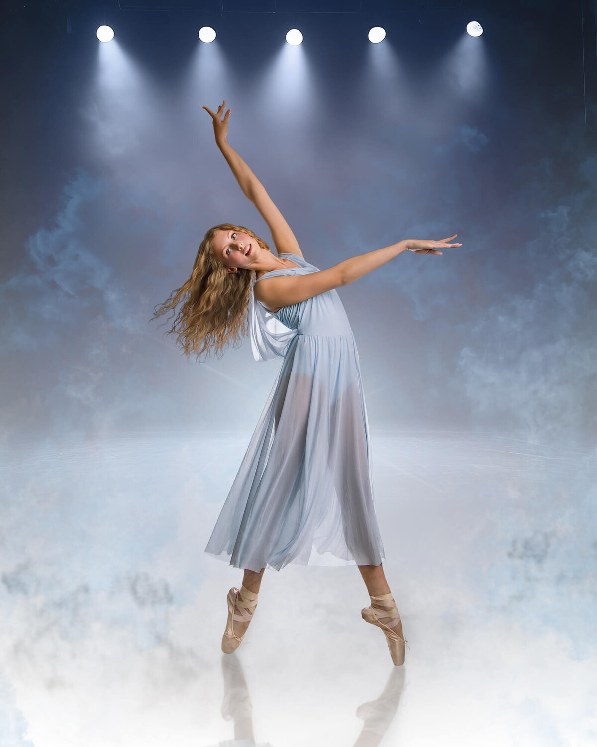 Beautiful blonde Ballerina in a graceful pose wearing a light blue sheer dress on a smokey blue stage