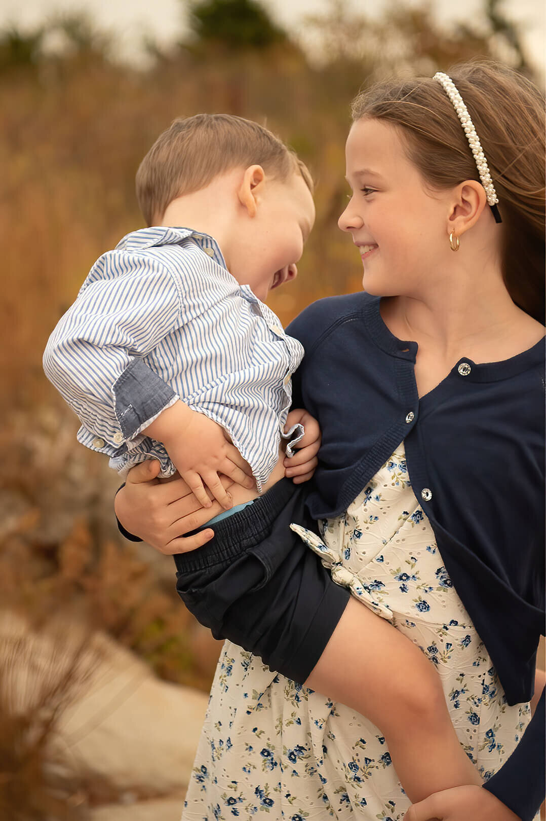 A big sister holds her little brother on her hip while he laughs at Wingaersheek Beach.