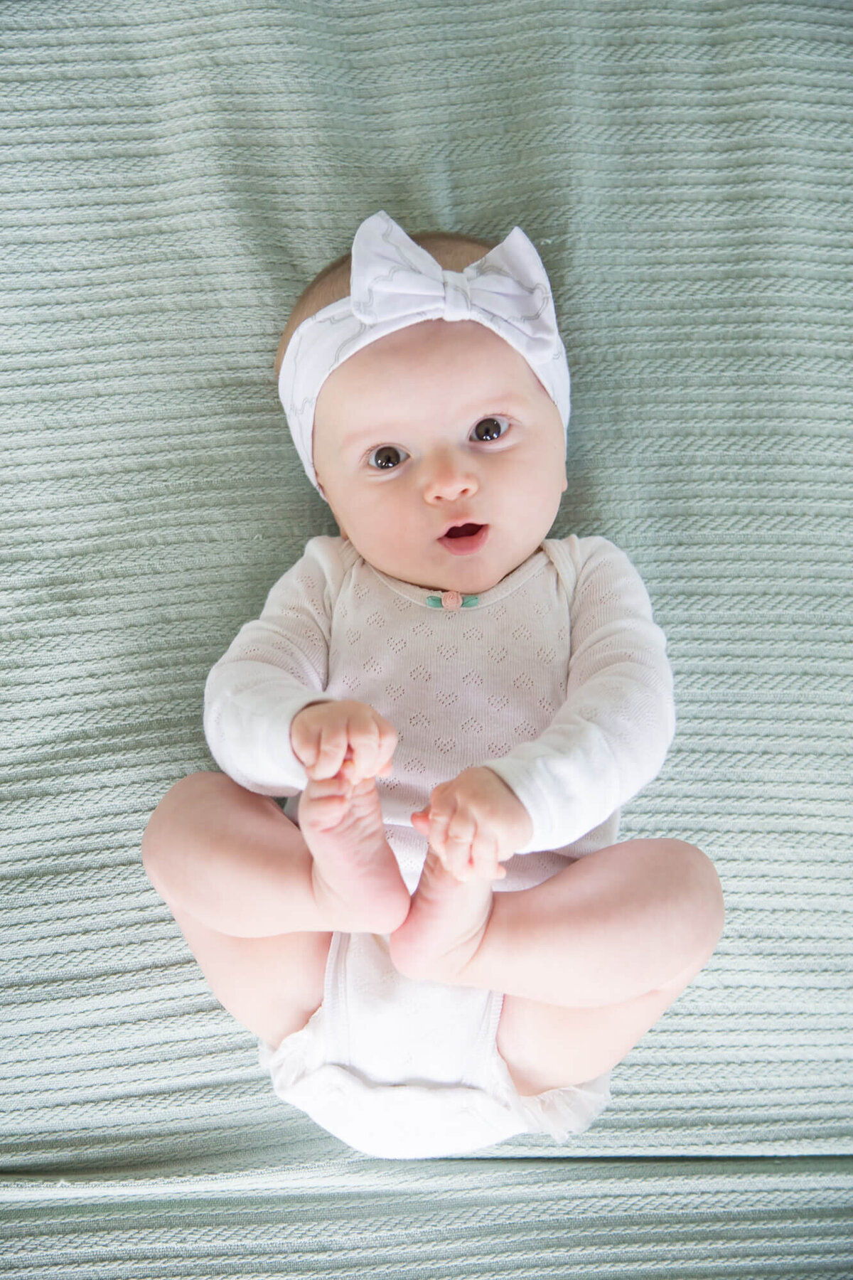 View from above of infant baby girl holding her toes and laying on a bed