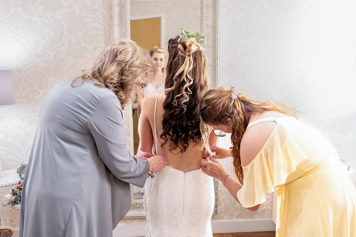 The bride stands in front of a mirror as two women help her zip up her wedding dress, capturing a sweet and emotional behind-the-scenes moment before the ceremony