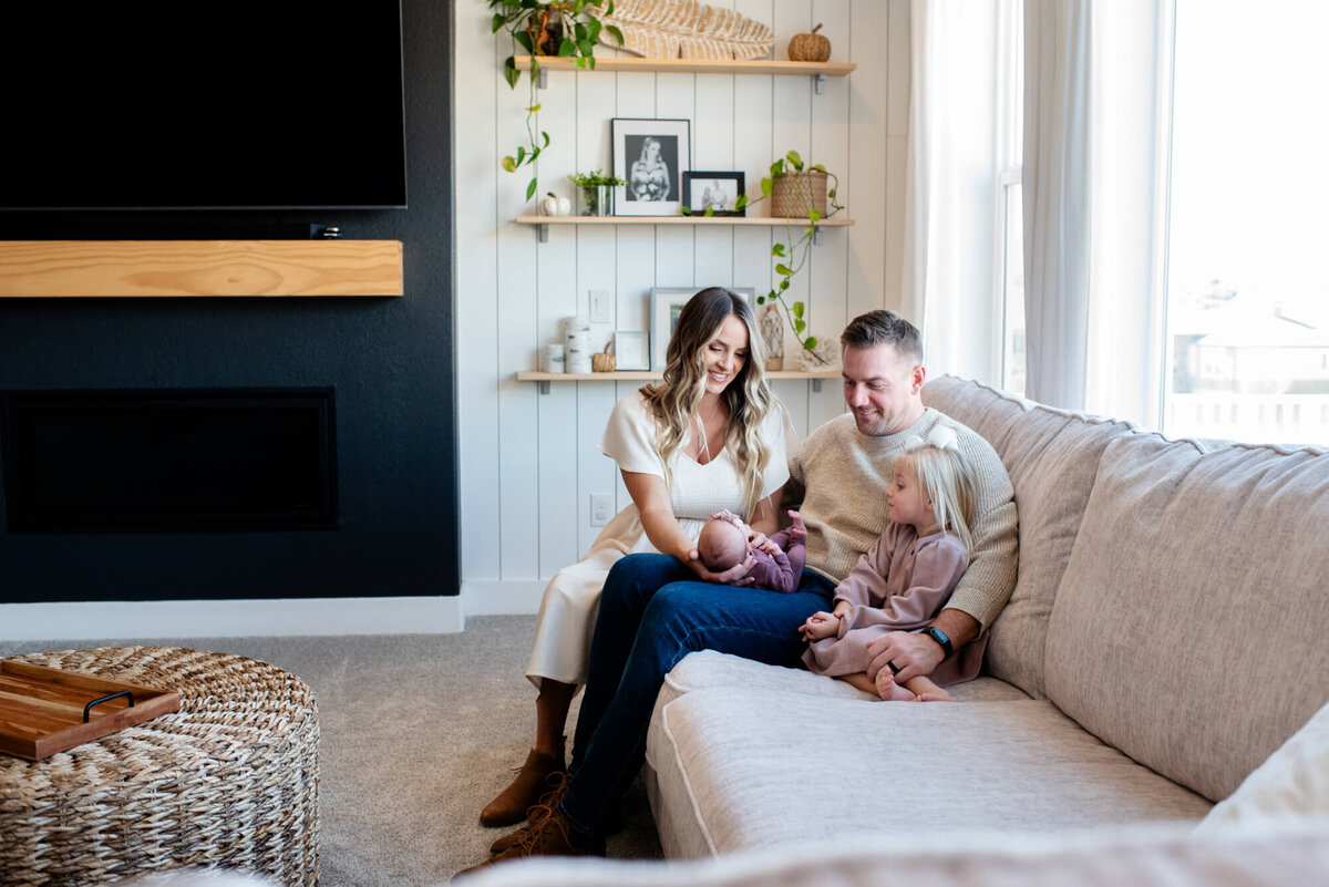 A mom and dad laugh while sitting on a couch with their newborn and toddler daughter