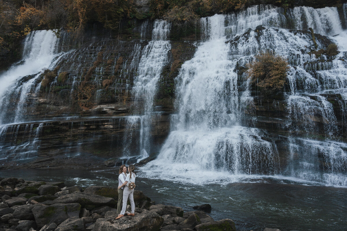At a beautiful waterfall in Tennessee, two brides dressed in suspenders embrace each other in celebration of their love during their elopement 