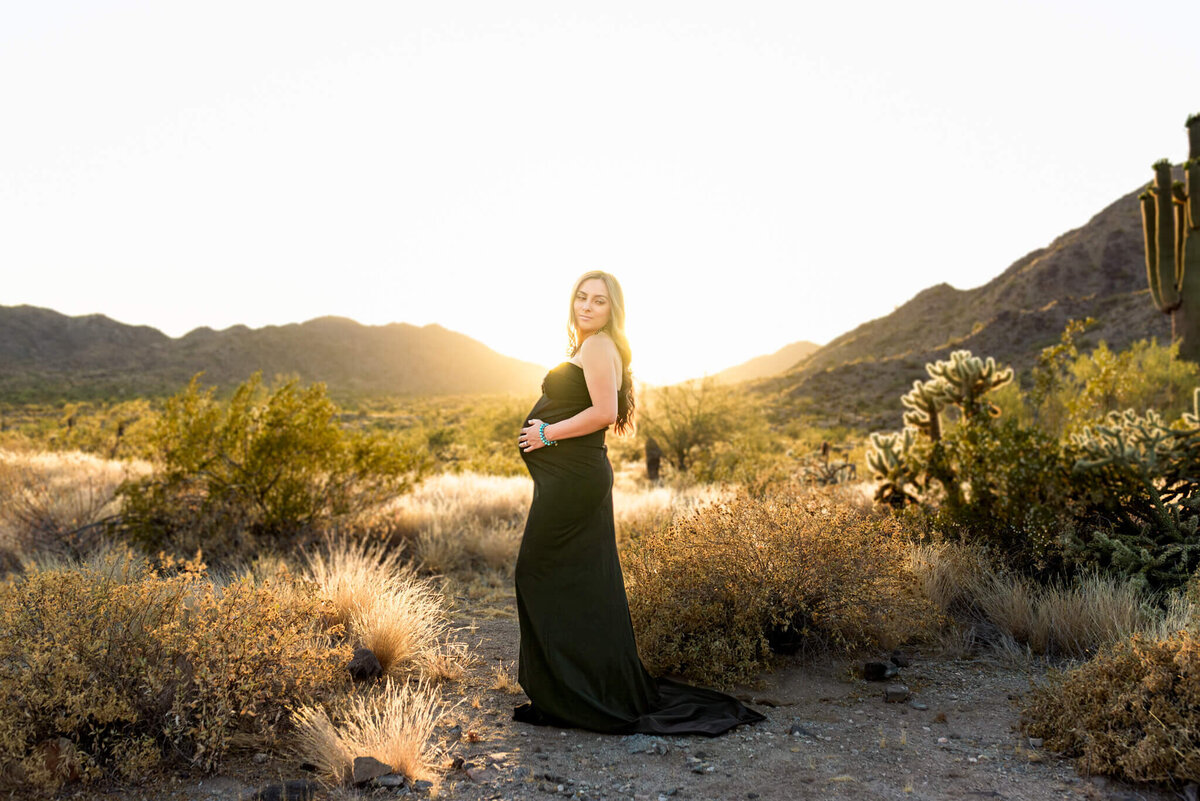 Glowing mom-to-be wearing fitted black gown at sunset in Arizona desert. Captured by Cactus & Pine Photography