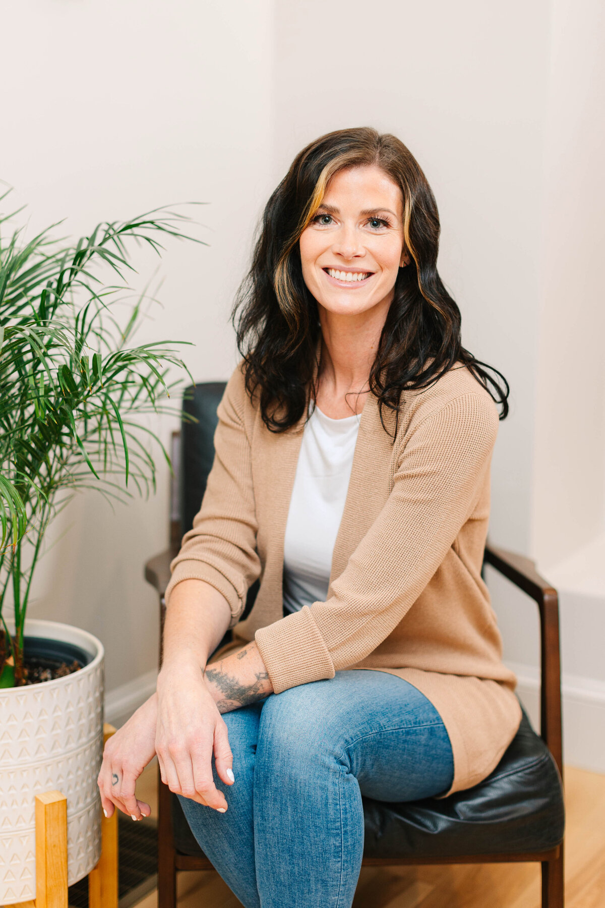 A smiling woman with wavy dark hair and blonde highlights sits in a modern black leather chair, wearing a beige cardigan, white top, and blue jeans. A potted plant is beside her in a neutral-toned room