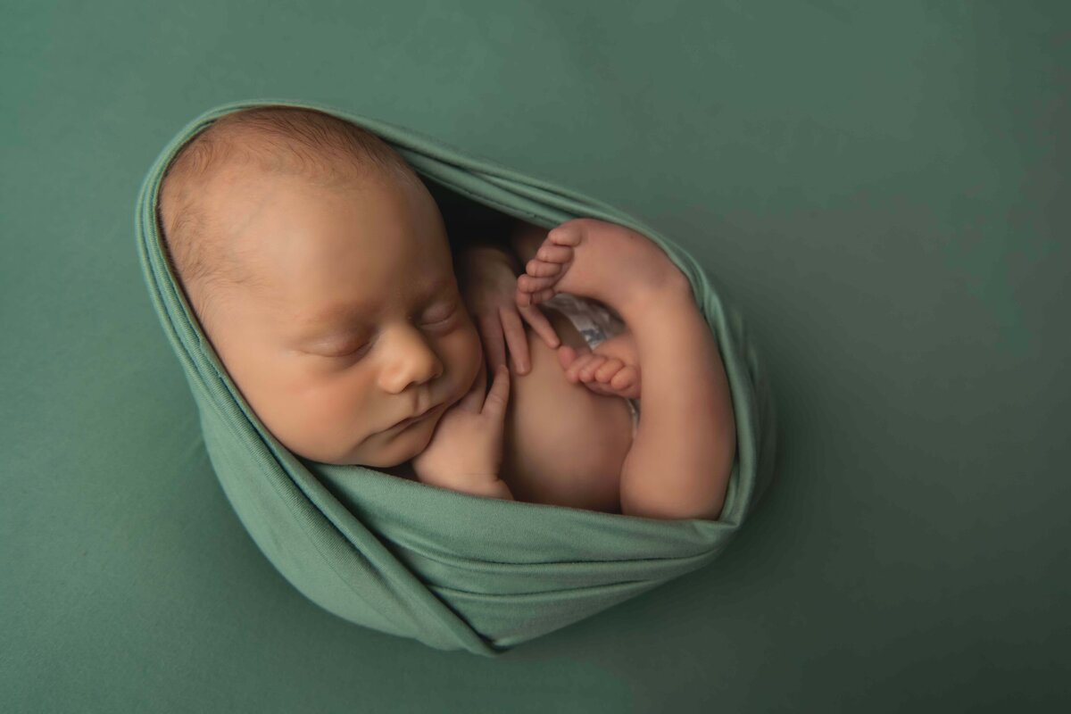 A newborn boy is posed carefully in this beautiful newborn photo done by Mobile, AL Newborn Photographer. The boy is curled up in a ball, as if he is still in the womb. He is wrapped in a teal blanket and is laying on a matching teal backdrop.