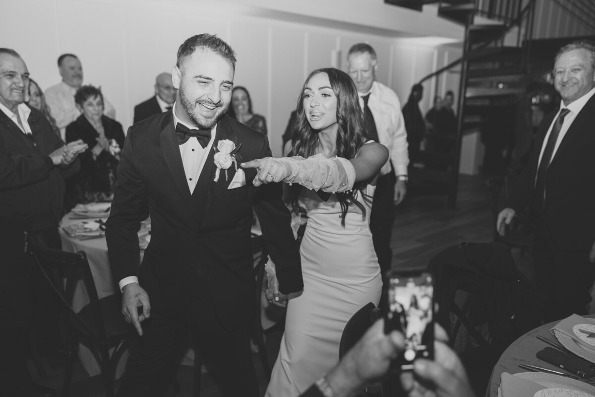 bride and groom walking into wedding reception smiling while bride points into crowd