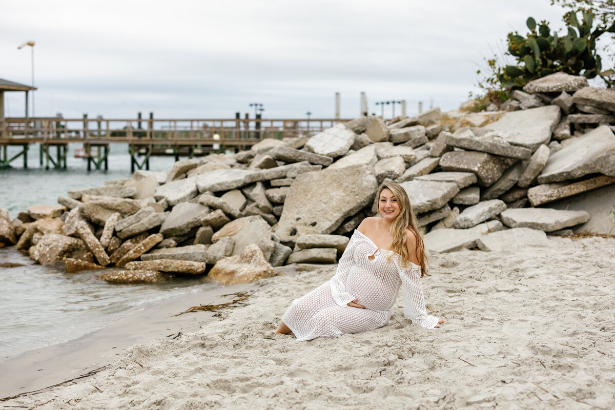 Pregnant woman cradling her bump while sitting on Wilmington beach sand, waves in background