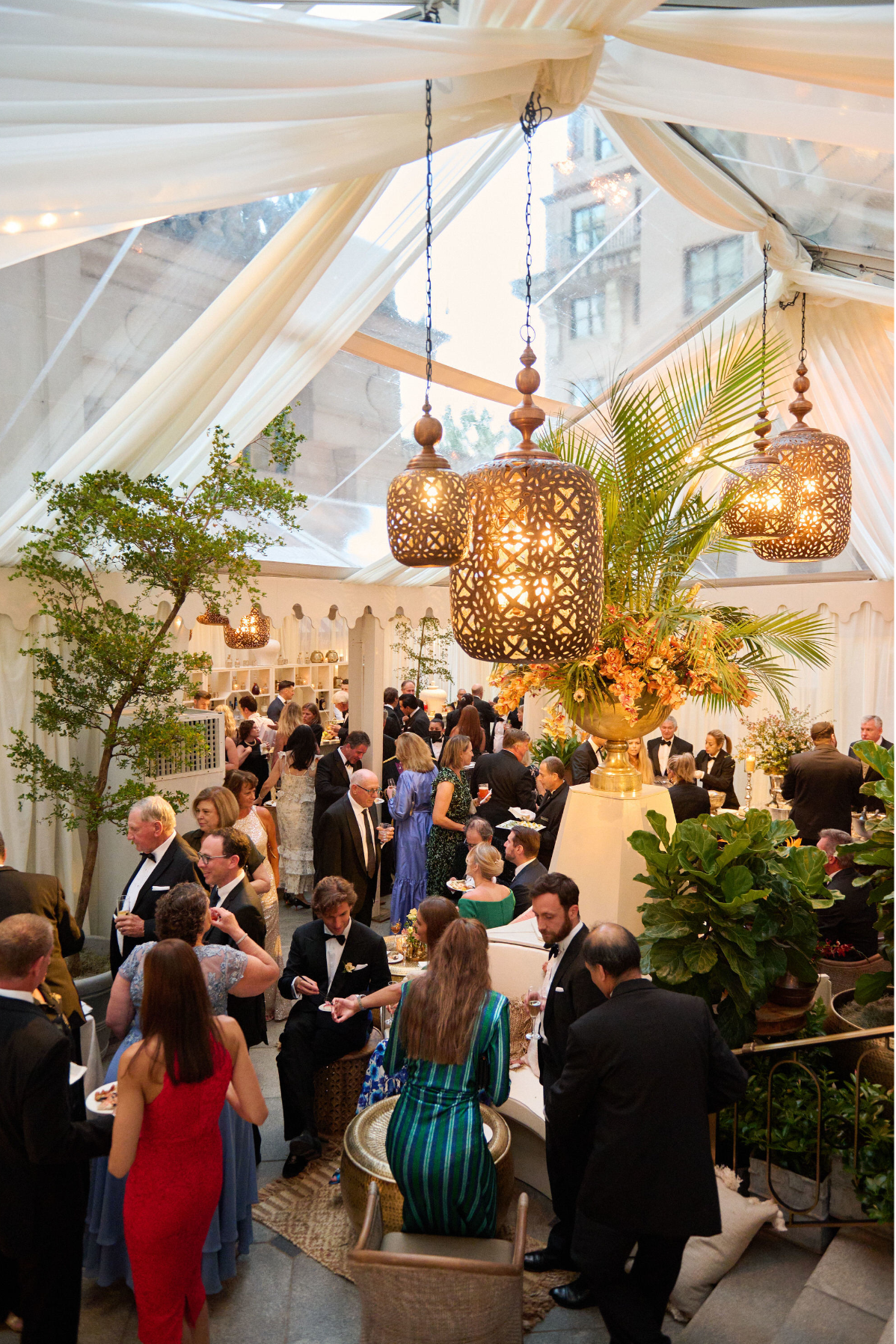 The photo was taken at Washington D.C., specifically at The Willard Intercontinental, one of the top wedding venues and hotels in DC, captured by photographer Shawn Connell. The scene depicts a lively indoor gathering, with elegantly dressed people engaging in conversation. The venue is adorned with lush green plants, elegant hanging lamps, and white drapery, creating an atmosphere of sophistication and elegance. Guests are seen standing around tables adorned with an array of delicious food and drinks under a semi-transparent canopy, adding to the ambiance of the event. Clayton Films provided videography services to capture the special moments of the occasion.